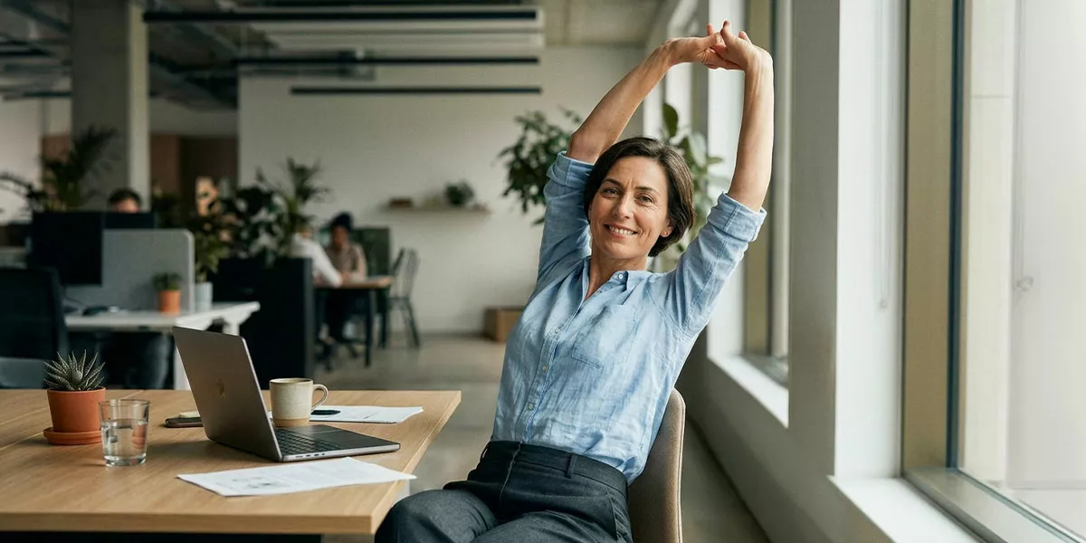 Eine lächelnde Frau streckt sich an ihrem Schreibtisch in einem modernen Großraumbüro