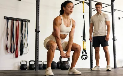 Eine Frau trainiert mit einer Kettlebell unter Anleitung ihres Trainers in einem hellen Studio