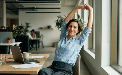 Eine lächelnde Frau streckt sich an ihrem Schreibtisch in einem modernen Großraumbüro