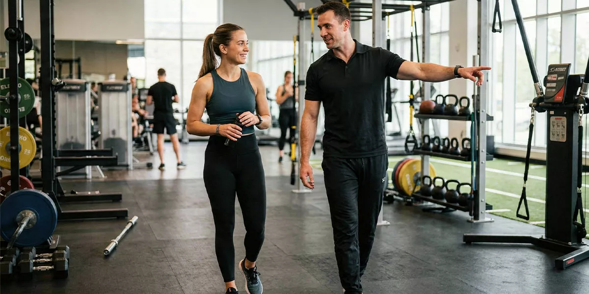 Ein Trainer und eine Trainerin laufen durch ein Fitnessstudio mit großer Fensterfront